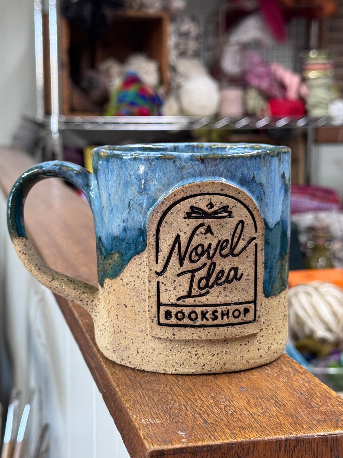 A handmade ceramic mug with a blue and beige glaze sits on a wooden shelf in a shop setting. The front of the mug features a raised label design that reads 'A Novel Idea Bookshop'.