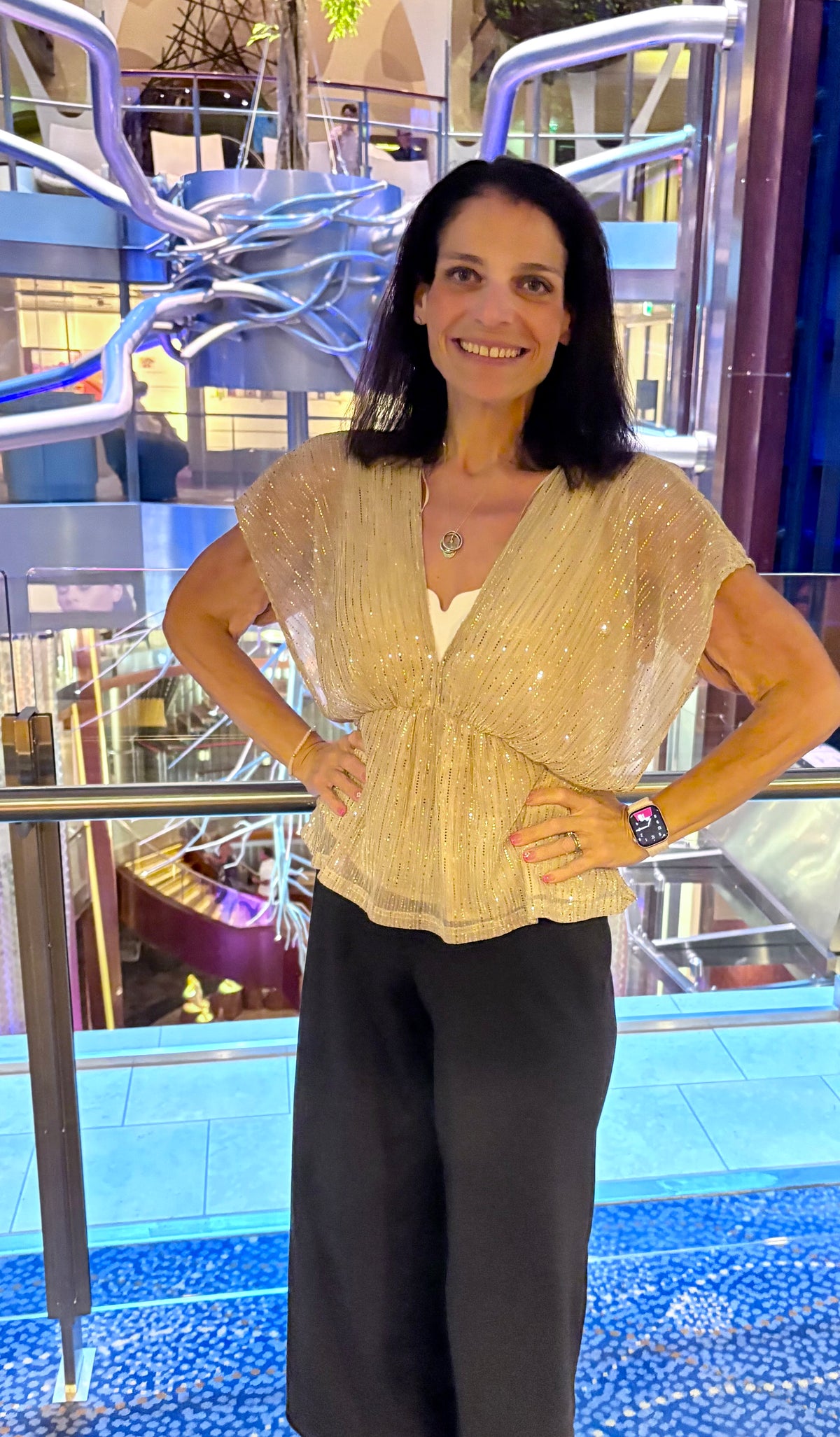 A woman posing in a shimmering champagne-colored sequined top and black wide-leg pants, standing in front of a glass railing with a blue patterned floor and a modern indoor resort setting in the background.