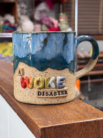 A handmade ceramic mug with a blue and teal glazed top half and a speckled beige bottom half. The mug features raised 3D lettering that reads 'A WOKE DISASTER' in various colors, sitting on a wooden shelf with a blurred shop background.