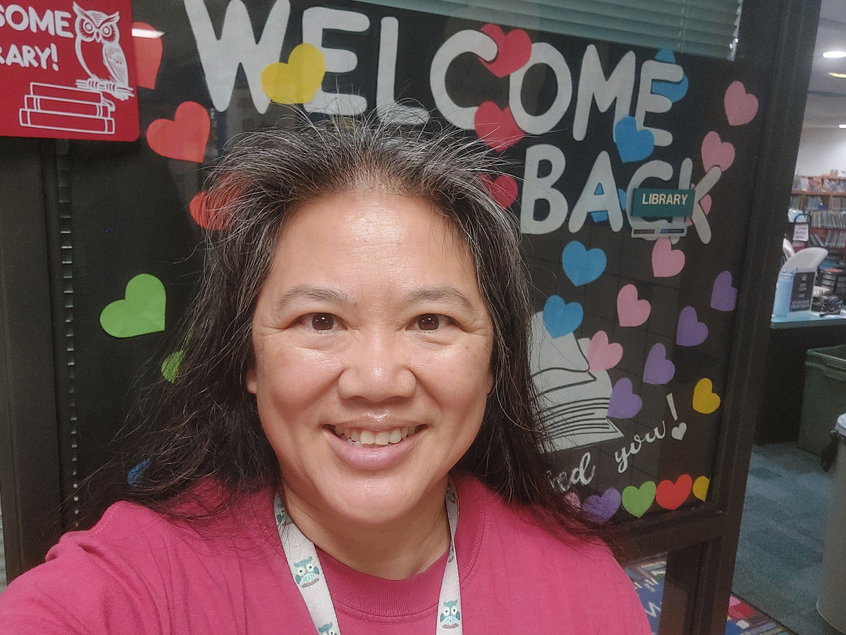 A smiling woman with dark hair wearing a pink top takes a selfie in front of a library welcome sign decorated with colorful hearts and the words 'WELCOME BACK' and 'LIBRARY'.