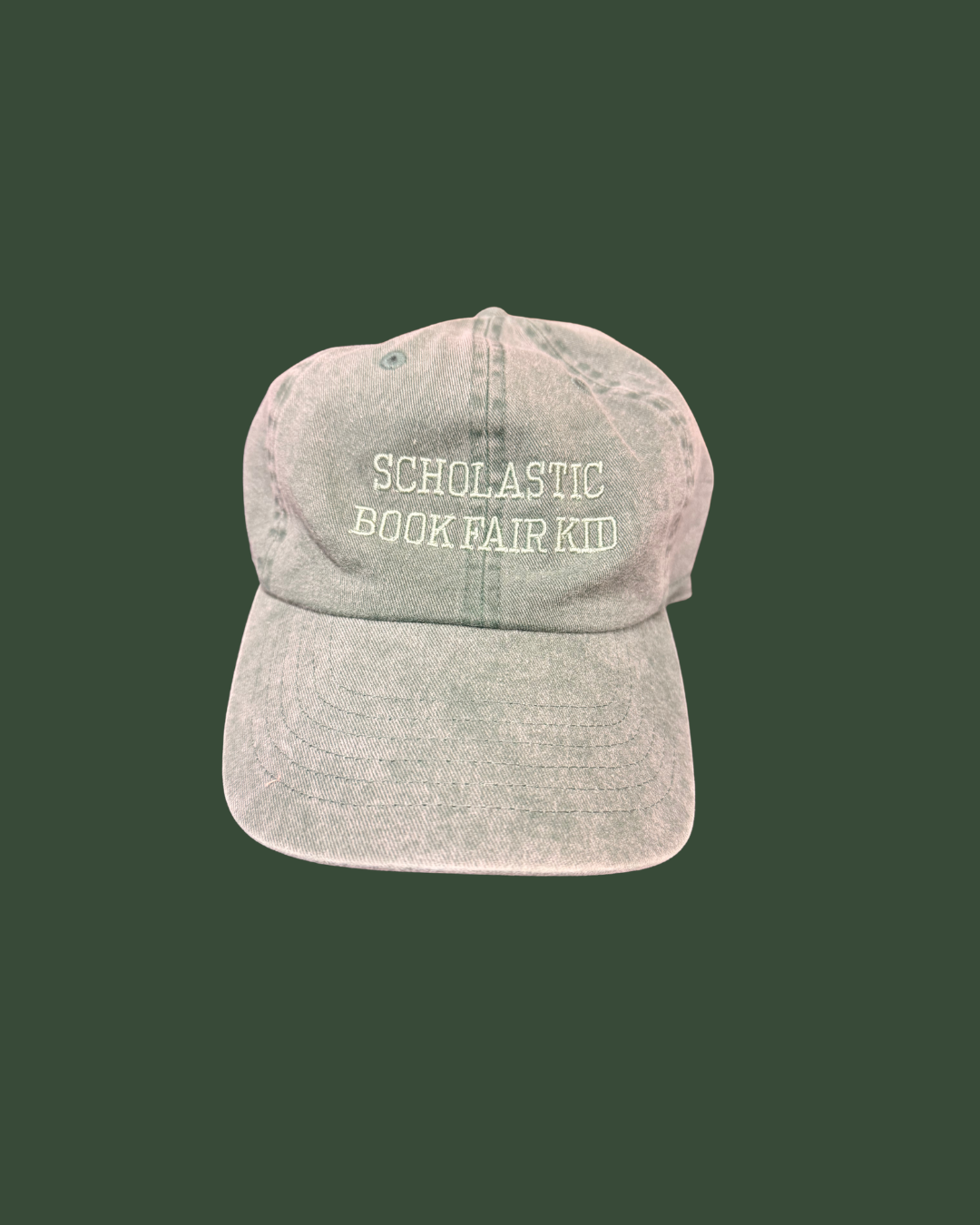 A light gray greenish acid-wash baseball cap with 'SCHOLASTIC BOOKFAIR KID' embroidered in white on the front, set against a solid dark green background.