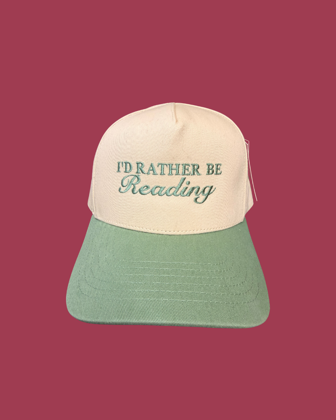A two-tone baseball cap with an off-white crown and a sage green brim, featuring the embroidered text 'I'D RATHER BE Reading' in green on the front. The cap is displayed against a solid dark pink background.