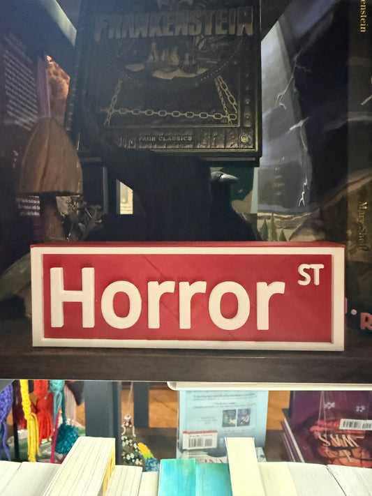 A red rectangular sign with white text reading 'Horror ST' displayed on a shelf in a store or room setting, with books and framed art visible in the background.