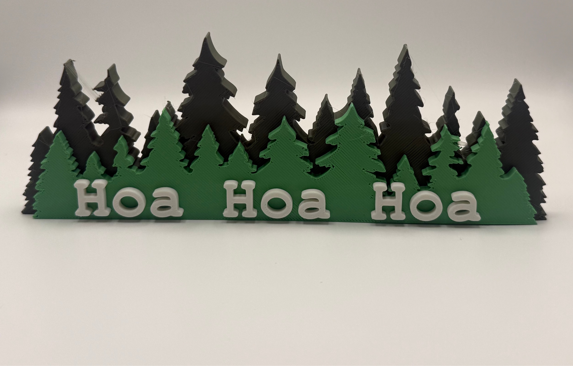 A small decorative sign featuring layered green and black pine tree silhouettes with the word 'Hoa Hoa Hoa' in white letters across the front, photographed on a plain light background.