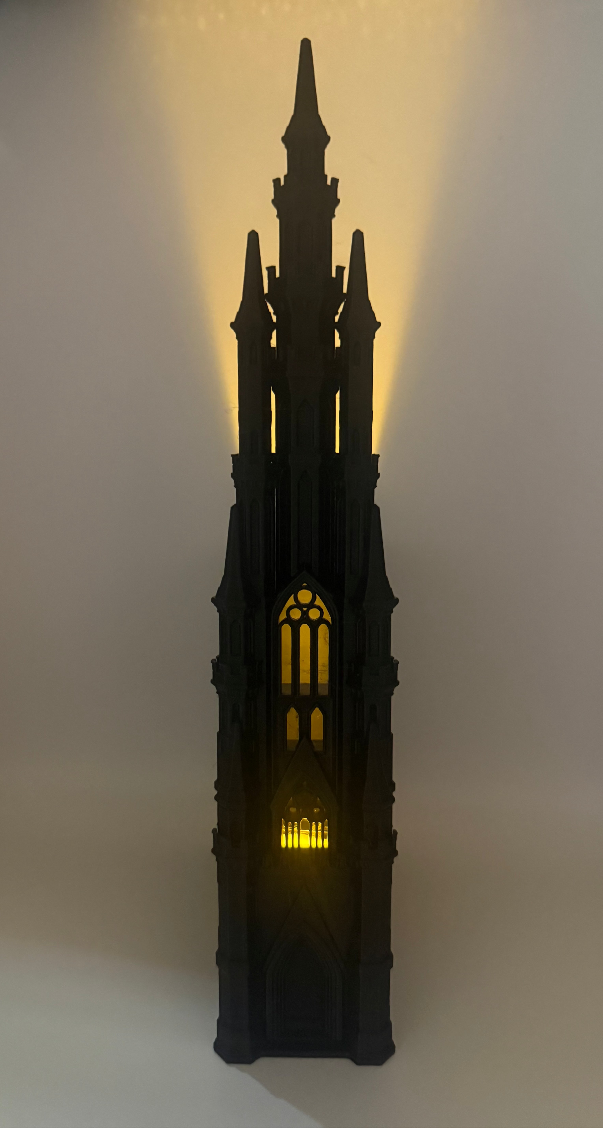 A black gothic cathedral tower figurine or lamp with warm yellow light glowing from within, set against a plain light background.
