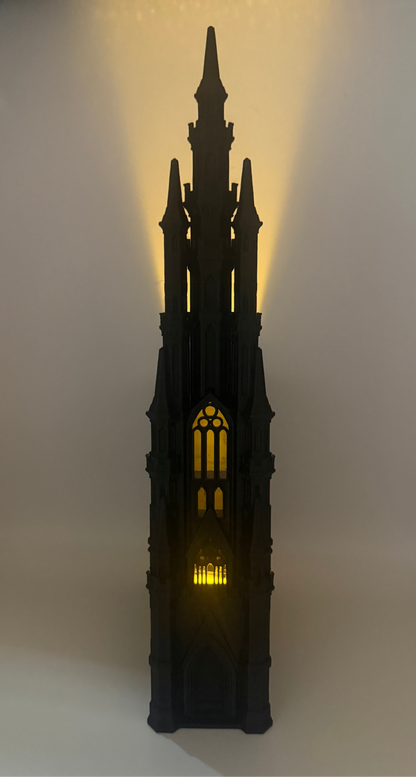 A black gothic cathedral tower figurine or lamp with warm yellow light glowing from within, set against a plain light background.