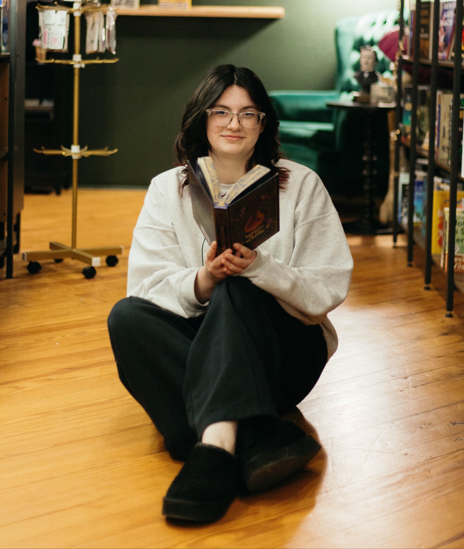 A person sitting on the floor in a cozy bookshop aisle, holding a book. The shop has tall bookshelves on both sides, a green wall with a 'Book Club' sign, wooden shelves, and a teal chair in the background.