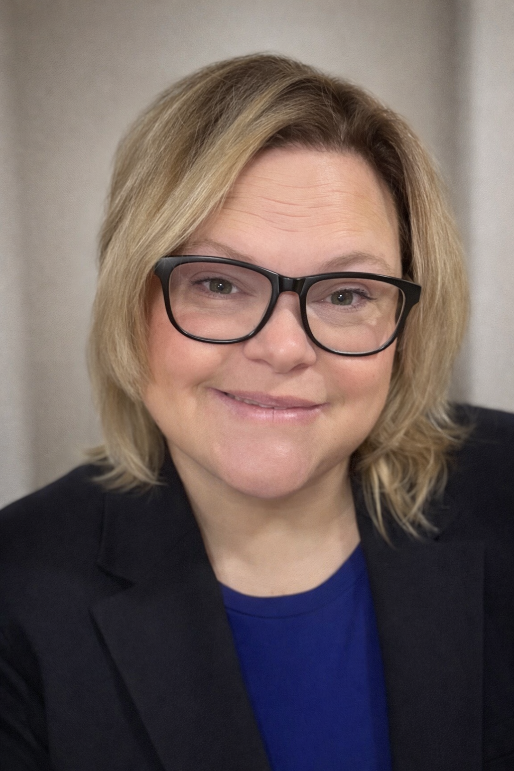 A professional headshot of a woman with blonde hair and black-rimmed glasses, wearing a black blazer over a royal blue top, set against a neutral beige background.