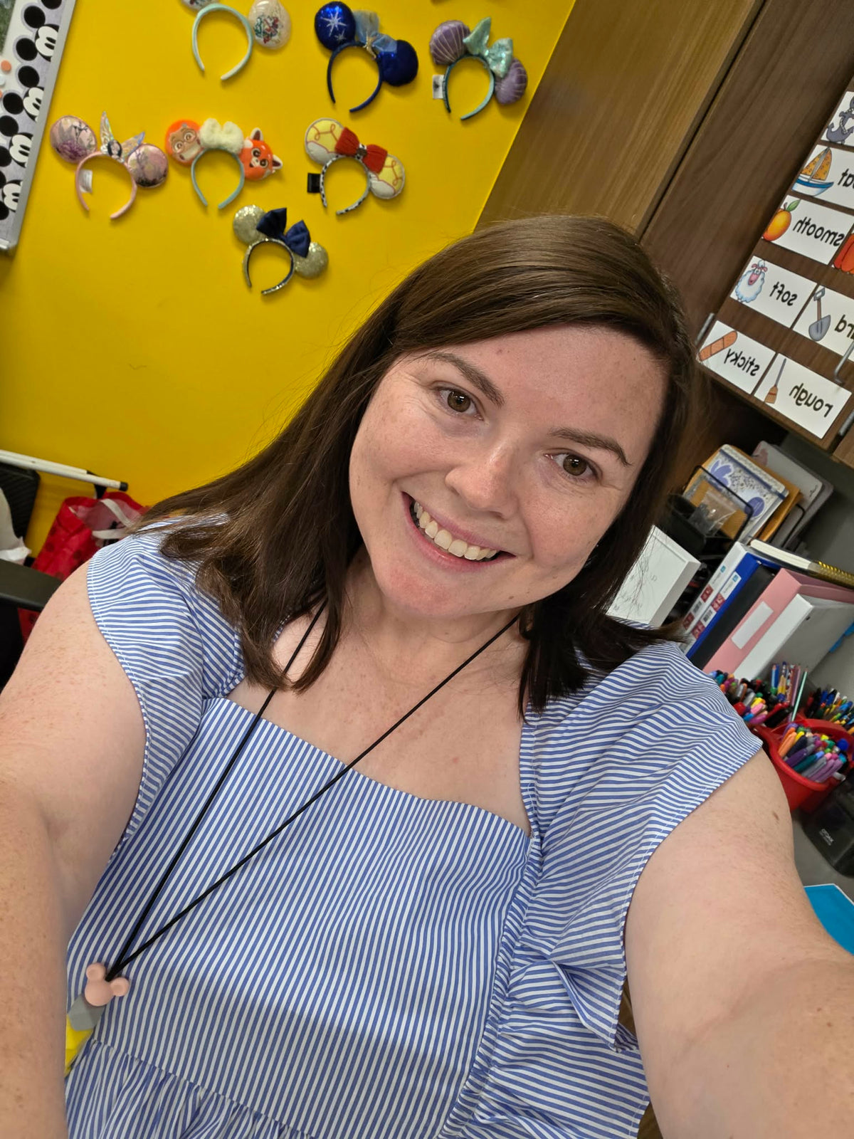 A smiling woman with brown hair takes a selfie in a classroom or office setting. She is wearing a blue and white striped sleeveless top.