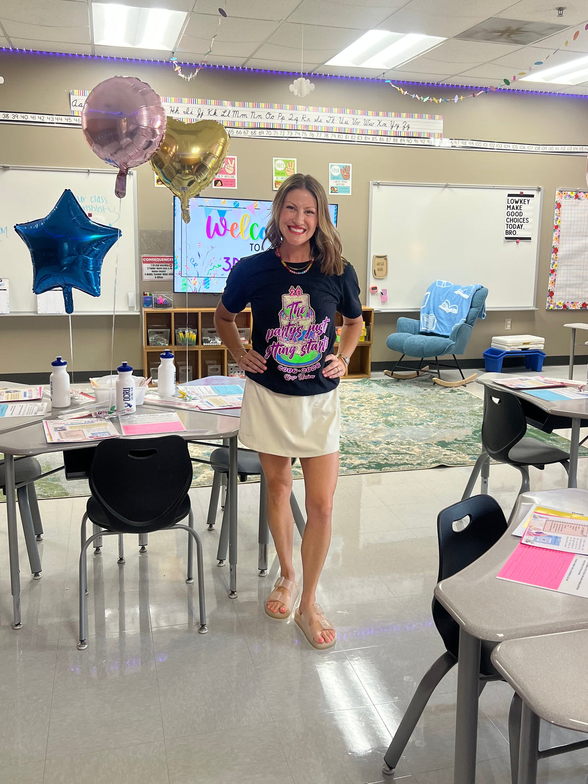 A woman stands in a decorated classroom wearing a dark navy graphic t-shirt with a pink and green cake design, paired with a cream-colored mini skirt. The room features balloons, colorful banners, and school desks.