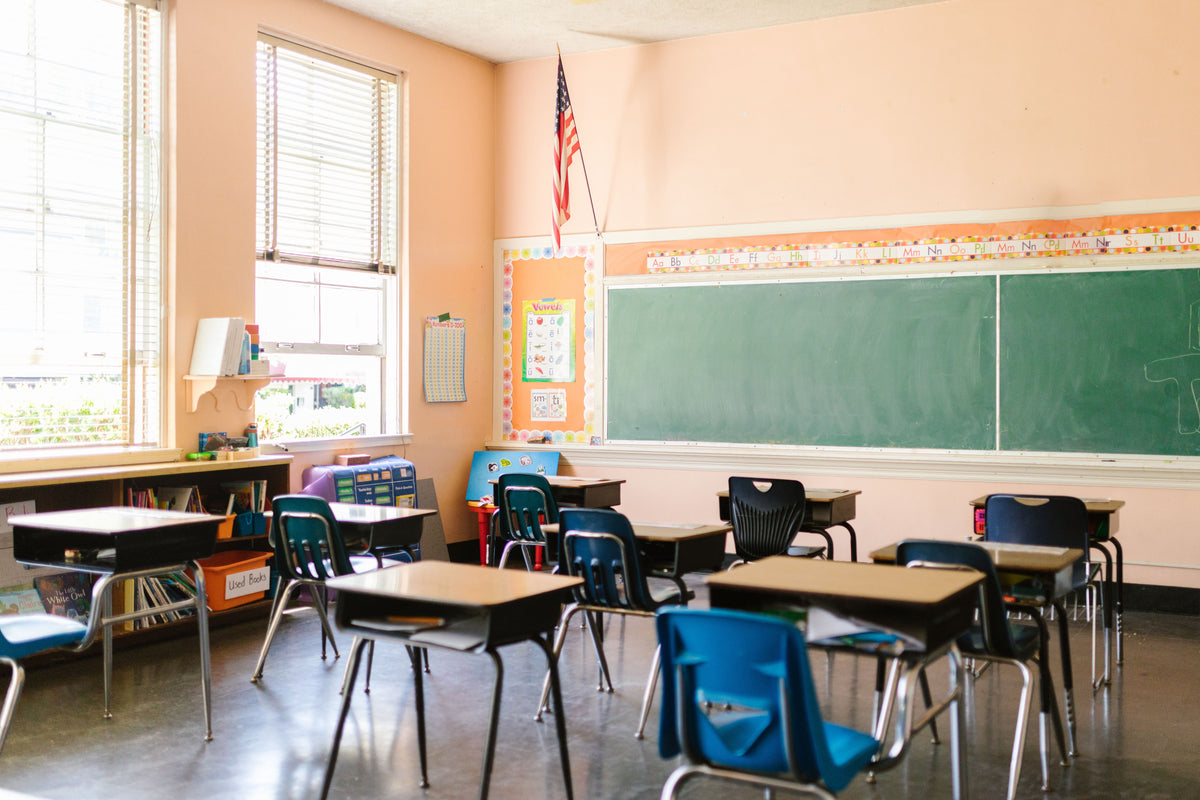 A bright classroom with rows of small desks and chairs, a large green chalkboard on the wall, peach-colored walls, windows with blinds, and an American flag hanging near the front.