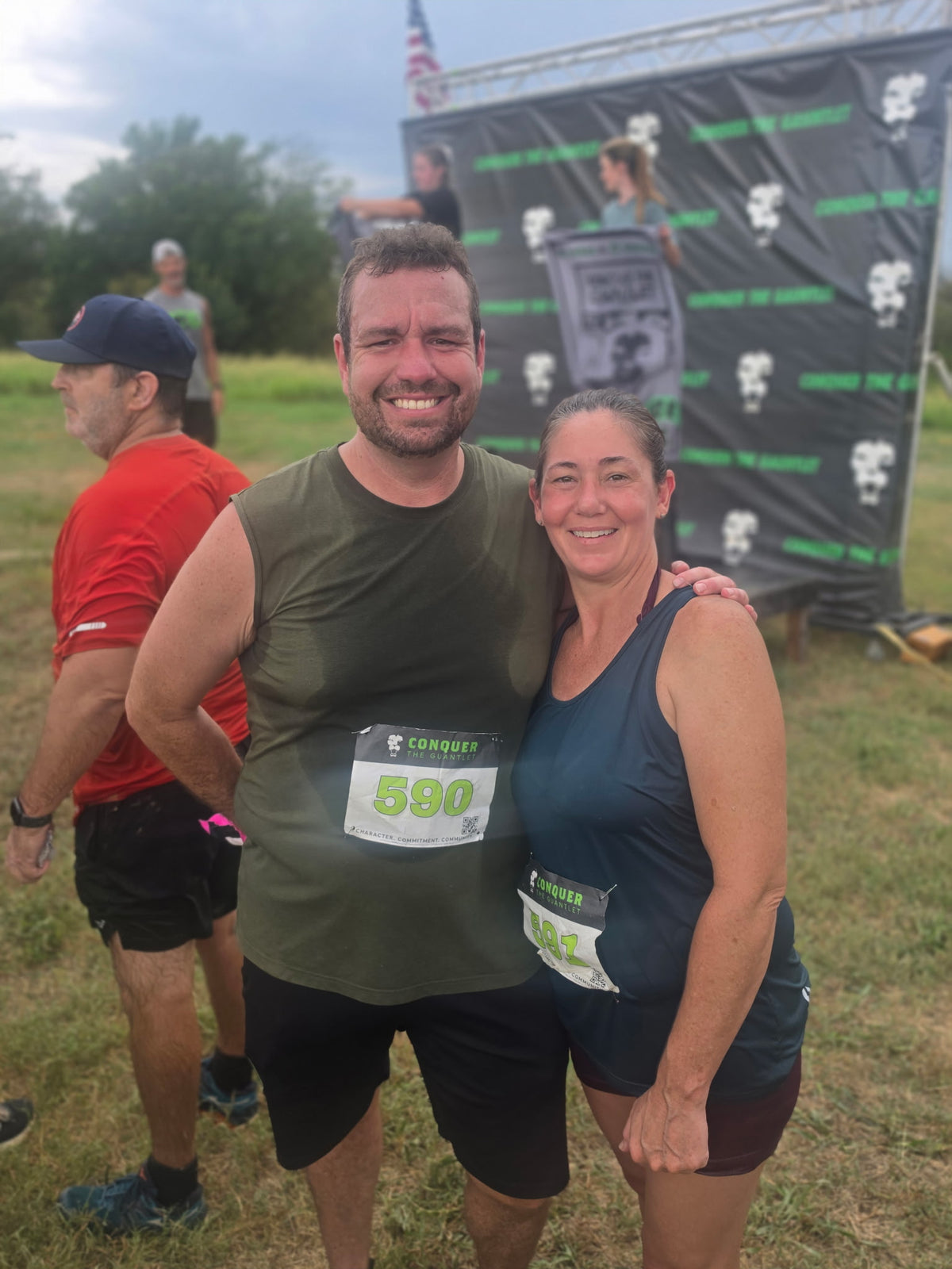 Two smiling race participants posing together outdoors at a race, with a black branded backdrop and grassy field in the background. The man wears an olive green tank top and black shorts, and the woman wears a dark blue tank top and dark shorts.